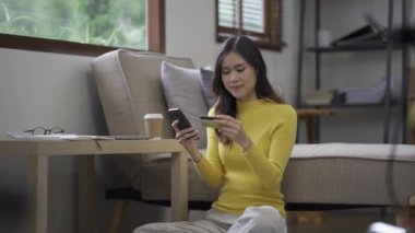 Kanepede otururken, elinde online alışveriş için kredi kartı tutan Asyalı bir kadın akıllı telefon kullanıyor ve evde internet üzerinden alışveriş yapıyor.