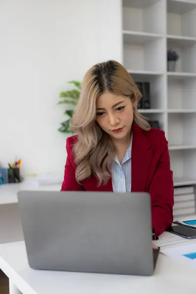 Asian female financial analyst or accountant uses laptop to work on financial audit and budgeting using calculator, checking balance sheet, working with company quarterly report. Vertical image