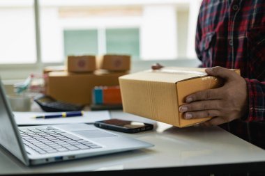 Yakın plan: Küçük bir KOBİ 'yi çalıştıran genç bir adam dizüstü bilgisayar ve hesap makinesi kullanıyor. KOBİ paketleme kutusunu incelemek için, e-ticaret ekibi konsepti ürünleri İnternet üzerinden satıp ev teslimatı sağlıyor.