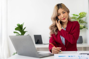 Ofiste dizüstü bilgisayarla ve finansal belgelerle çalışırken iş müşterisiyle telefonda konuşan çekici Asyalı kadın muhasebeci.