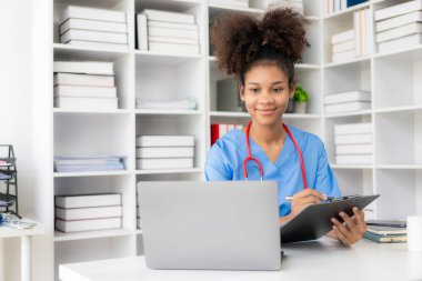 Beyaz önlüklü ve steteskoplu başarılı Amerikalı kadın doktor hastanede dizüstü bilgisayarla çalışıyor, hastalara danışmanlık yapıyor..