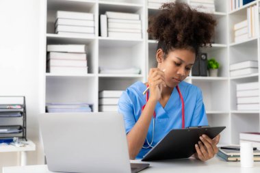 Beyaz önlüklü ve steteskoplu başarılı Amerikalı kadın doktor hastanede dizüstü bilgisayarla çalışıyor, hastalara danışmanlık yapıyor..