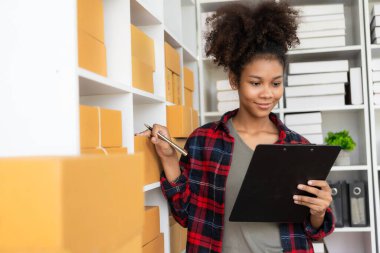 Evden çalışan Afro-Amerikalı bir kadın KOBİ paketlerini küçük işletme ofisinde teslimat için hazırlıyor. Laptop bilgisayarı üzerinde çalışan bir kadın. Posta paketiyle internette fikirler satıyor.