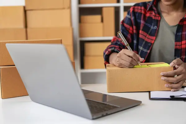 African American woman working from home preparing SME parcel box for delivery at small business office, woman working on laptop computer from home on top with postal parcel selling ideas online