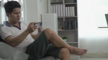 Evdeki oturma odasında kanepede otururken cep telefonu kullanan mutlu Asyalı genç adam. Ve evdeki cep telefonundan konuş ve rahatla. Futbol maçlarını izlemenin tadını çıkar.