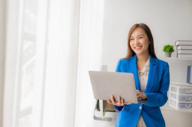 Asyalı kız elinde dizüstü bilgisayarla ofiste çalışıyor, aynalı pencerenin yanında duruyor, online çalışma kavramı