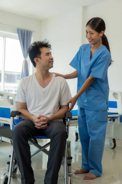 Genç bir bayan doktor evde tekerlekli sandalyedeki bir adamla ilgileniyor. Pozitif ve mutlu Asyalı kadın hemşire ya da mavi üniformalı fizyoterapist yakın çevredeki erkek hastayla ilgileniyor..