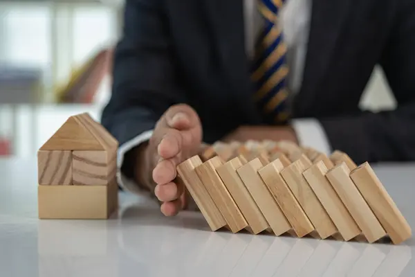 Cropped view of risk manager protecting house model from falling wooden blocks with hands, home insurance and safety. Businessman\'s hands stop wooden blocks from falling onto the model house.