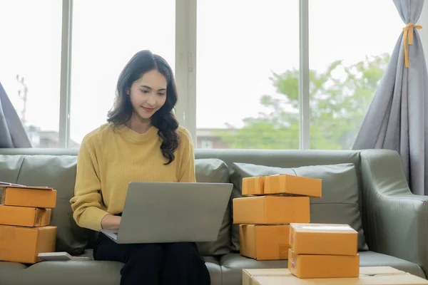 Asian woman working from home Online sales storage, e-commerce Business owners use laptops or tablets to receive and verify online orders to prepare boxes of products. online shopping