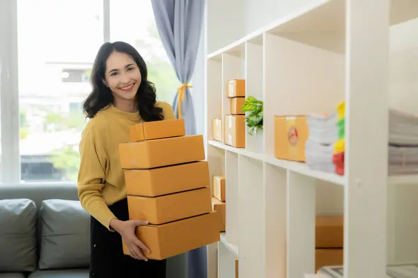 Asian woman working from home Online sales storage, e-commerce Business owners use laptops or tablets to receive and verify online orders to prepare boxes of products. online shopping