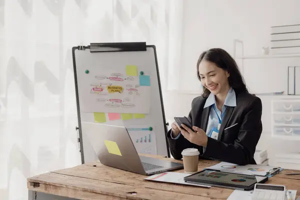 Happy young Asian businesswoman or sales manager wearing suit working in contemporary glass office talking with customers on phone using laptop at work, finance business concept