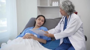 Kıdemli Asyalı kadın doktor klinikte hasta yatağında yatan genç Asyalı kadına danışmanlık yapıyor, muayene sonuçlarını, sağlık tavsiyelerini veya internetteki reçeteyi tablet bilgisayardan 4k 60p ile tartışıyor.