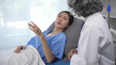 Kıdemli Asyalı kadın doktor klinikte hasta yatağında yatan genç Asyalı kadına danışmanlık yapıyor, muayene sonuçlarını, sağlık tavsiyelerini veya internetteki reçeteyi tablet bilgisayardan 4k 60p ile tartışıyor.