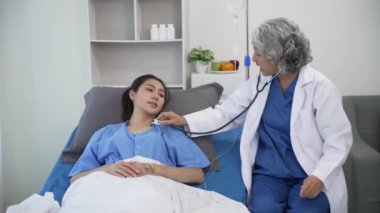 Kıdemli Asyalı kadın doktor klinikte hasta yatağında yatan genç Asyalı kadına danışmanlık yapıyor, muayene sonuçlarını, sağlık tavsiyelerini veya internetteki reçeteyi tablet bilgisayardan 4k 60p ile tartışıyor.