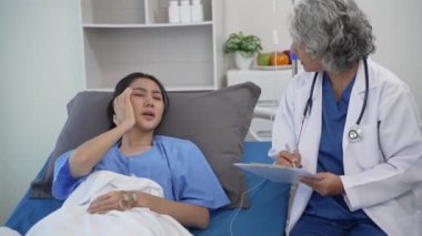 Kıdemli Asyalı kadın doktor klinikte hasta yatağında yatan genç Asyalı kadına danışmanlık yapıyor, muayene sonuçlarını, sağlık tavsiyelerini veya internetteki reçeteyi tablet bilgisayardan 4k 60p ile tartışıyor.