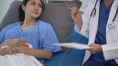 Kıdemli Asyalı kadın doktor klinikte hasta yatağında yatan genç Asyalı kadına danışmanlık yapıyor, muayene sonuçlarını, sağlık tavsiyelerini veya internetteki reçeteyi tablet bilgisayardan 4k 60p ile tartışıyor.