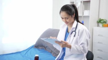 Beyaz cüppeli ve steteskoplu Asyalı kadın doktor bir hastanın yatağının yanında duran tıbbi not defterine not alıyor. Sağlık ve Tıbbi Konsept 4k 60p görüntü