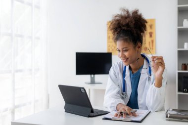 Beyaz önlüklü genç bir Afrikalı Amerikalı kadın doktor danışma masasında dizüstü bilgisayar kullanıyor. Doktor çevrimiçi video konferansı aracılığıyla hastayla sohbet ediyor.