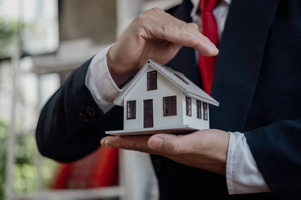 A businessman in a suit holds a house model in the palm of his hand. Mortgage Lending Concepts Real estate insurance and residential building sales. Close-up.