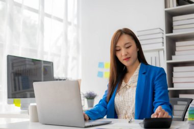 Genç Asyalı kadın bir masada oturuyor ve yatırım sonuçlarını gösteren finansal grafikleri hesaplıyor. İş büyüme sürecinizi hesap makinesi ve dizüstü bilgisayar kullanarak finansal inceleme ve bütçe ile planlayın.