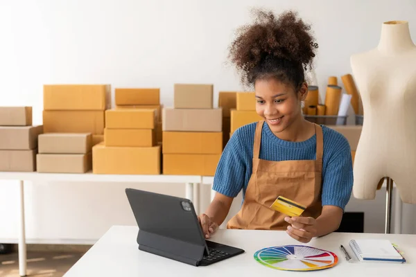 Young African woman running an online store, startup, small business, SME, using a smartphone or tablet, works with receipt boxes and checks online orders to prepare to pack boxes to sell to customer