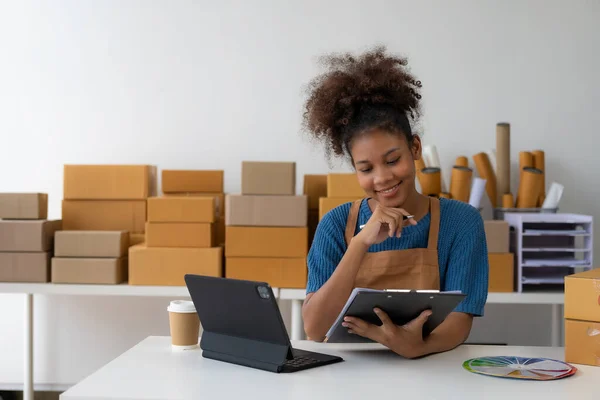 Young African woman running an online store, startup, small business, SME, using a smartphone or tablet, works with receipt boxes and checks online orders to prepare to pack boxes to sell to customer