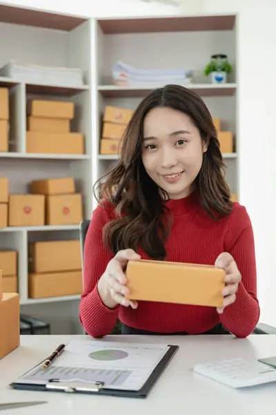 Kırmızı günlük elbiseli Asyalı kız gülümsüyor ve hazır ürünleri müşterilere teslim etmek için tutuyor. Ürün kutuları ve ambalaj ekipmanları ile, KOBİ iş konsepti evde çevrimiçi satış için.