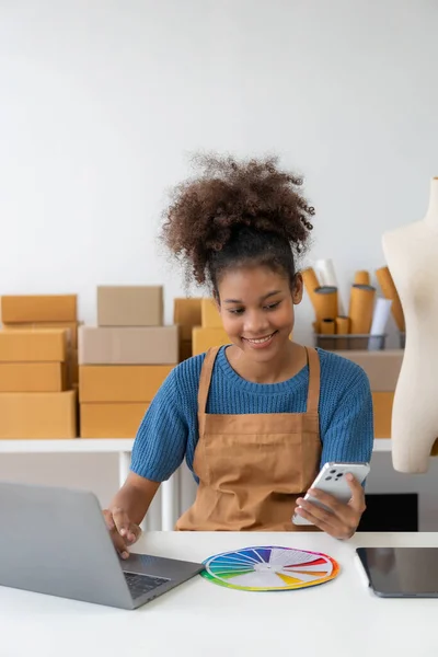 Startup, small business, African woman business owner using a smartphone or laptop to receive and verify online orders in order to prepare boxes of packed products for delivery. SME business concept