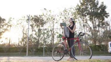 Asyalı kadın sırtında sırt çantasıyla parkta egzersiz yapmak için bisiklete biniyor. 4K video