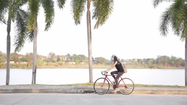アジアの女性は 彼女の背中のバックパックで公園で運動するために喜んで自転車に乗ります 4Kビデオ — ストック動画