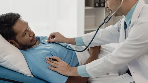 stock image Asian male doctor holding stethoscope and listening to patient Doctor doing heart check examining young man lying on bed, heart disease, health care concept.