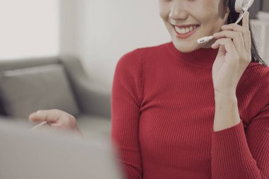 Çağrı merkezinde çalışan genç bir Asyalı kadın bilgisayar ve kulaklıkla mutlu bir şekilde gülümseyen ve bir müşteriyle sorunlarını çözmelerine yardımcı olmak için sevgi dolu bir konuşma yapan bir destek görevlisi..