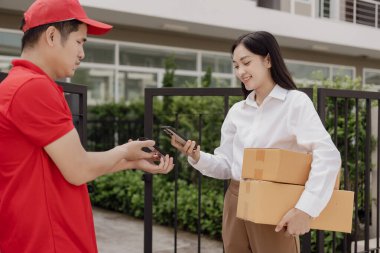 Genç Asyalı kadın, mutlu ve gülümseyen postacıdan bir karton kutu alır. Kurye, öğeyi acilen teslim eder. Çevrimiçi mağaza, kağıt konteynırlar, postacı, teslimat servisi, paketler