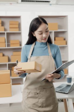 Posta paketi. Çevrimiçi satış. KOBİ başlangıç konsepti. Bağımsız Asyalı kadın küçük işletme girişimcisi. Önlük takıyor. İnternetten yemek sipariş etmek için laptop ve kutu kullanıyor. KME iş dikey resmi