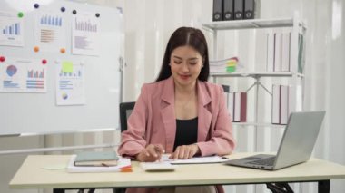 Finansal grafik belgeleriyle çalışan ve birikimleri hesaplamak için hesap makinesi kullanan genç Asyalı muhasebeci veya dizüstü bilgisayar ve vergi kopyalama, ticaret, araştırma, muhasebe belgesi, şirket 4k