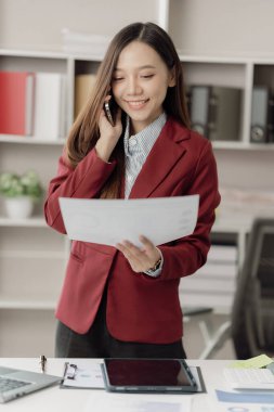 Ofiste dijital akıllı telefon kullanan, bilgisayarını kullanan, ofisteki mali belgelerle aynı telefonla konuşan mutlu Asyalı genç bir çalışan..