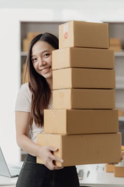 Önlük takan serbest Asyalı kadın, müşterilere satmak üzere paket hazırlamak için online siparişleri almak ve kontrol etmek için dizüstü bilgisayar ve kutu kullanıyor. KME çevrimiçi iş fikri, dikey resim