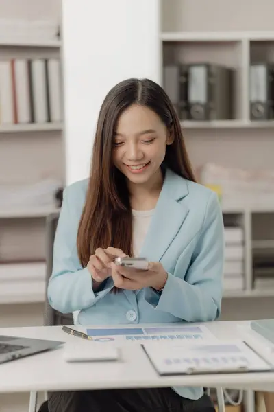 Happy young Asian employee using digital smartphone sitting in office work area, using laptop, talking on the same phone with financial documents in the office.