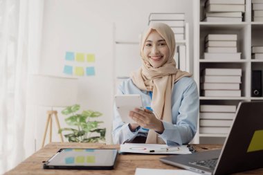 Asyalı Müslüman iş kadını hesap makinesi kullanır ve masadaki dizüstü bilgisayarlı ve mali belgeli, vergi, muhasebe, finansal hesaplı notlar yazar.