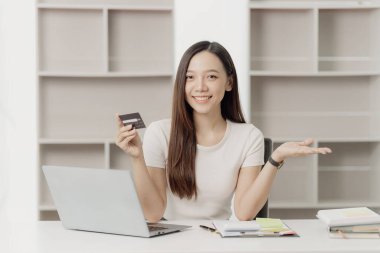 Gülümseyen genç bir kadın evde dizüstü bilgisayarla otururken, internetten alışveriş yaparken, kredi kartı tutarken, internet bankacılığı ve e-ticaret başvurularında ödeme yaparken kredi kartı tutuyor.