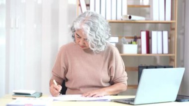 Yaşlı Asyalı kadın bilgisayar masasında evrak işleriyle uğraşırken evde ciddi yüz ve evrak yığını masada, ev işi konsepti.