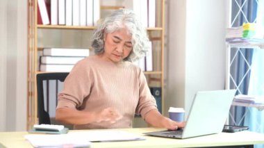 Yaşlı Asyalı kadın bilgisayar masasında evrak işleriyle uğraşırken evde ciddi yüz ve evrak yığını masada, ev işi konsepti.