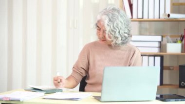 Yaşlı Asyalı kadın bilgisayar masasında evrak işleriyle uğraşırken evde ciddi yüz ve evrak yığını masada, ev işi konsepti.