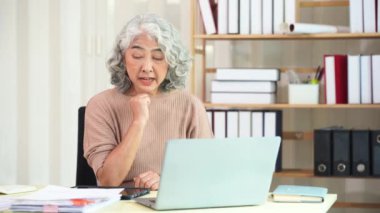 Yaşlı Asyalı kadın bilgisayar masasında evrak işleriyle uğraşırken evde ciddi yüz ve evrak yığını masada, ev işi konsepti.