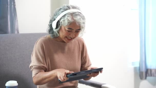 高齢アジアの女性は オンライン決済 ゲーム ソーシャルメディア チェックメールを書くためにタブレットデバイスを使用し リビングルームのソファに座って — ストック動画