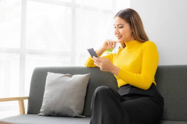 Sarı gömlek giyen kahverengi saçlı genç bir kadın kanepeye oturur, mesajlaşır ve gülümser. Mutlu model sosyal medyada eğlenceli bir video çağrısı yapıyor