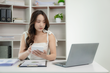 Genç Asyalı mutlu bir çalışan masada oturuyor ve bilgisayar ve hesap makinesiyle çalışıyor. Mali belgeler, hukuk şirketinin vergi oranlarını hesaplıyor..