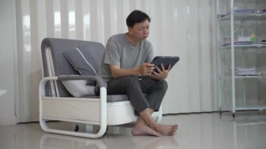 Kanepe üzerinde tablet kullanan gülümseyen Asyalı yaşlı bir adam tablet kullanıyordu. Gözlüklü neşeli yaşlı bir adam internette sohbet ederken eğleniyordu. Evdeki internete bağlan