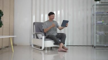 Kanepe üzerinde tablet kullanan gülümseyen Asyalı yaşlı bir adam tablet kullanıyordu. Gözlüklü neşeli yaşlı bir adam internette sohbet ederken eğleniyordu. Evdeki internete bağlan