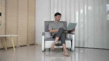 Yaşlı Asyalı adam dizüstü bilgisayarını kullanarak iyi vakit geçiriyor, sosyal medyayı kontrol ediyor, haberleri okuyor ve evdeki oturma odasındaki kanepede otururken internetten alışveriş yapıyor..